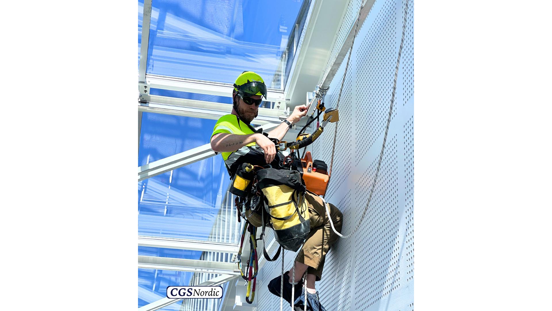 CGS Nordic technician installing H-RAIL rigid rail track on steel structure under glazed roof, using professional power tools during installation of rope access system.