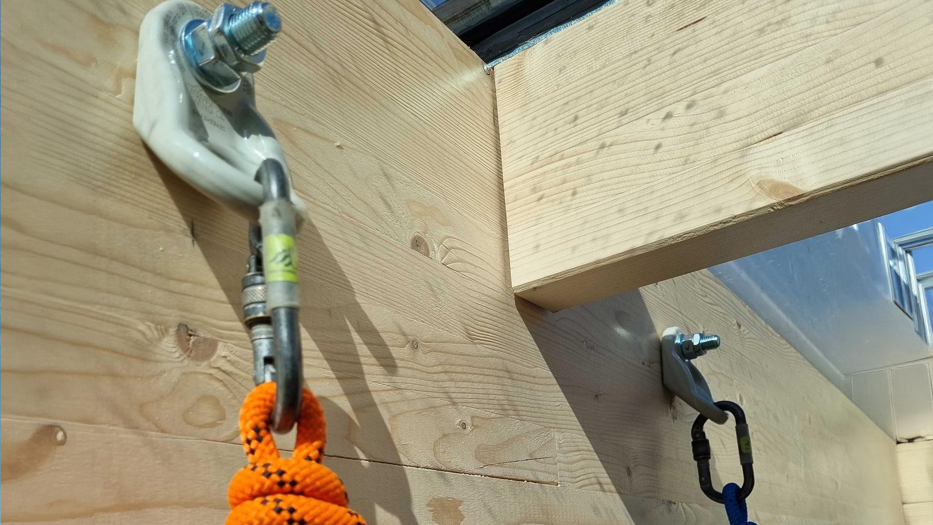 Rothoblaas WING single anchor points installed on a glulam timber beam junction — rope access carabiners and ropes connected for suspended work below a timber roof structure, Denmark.