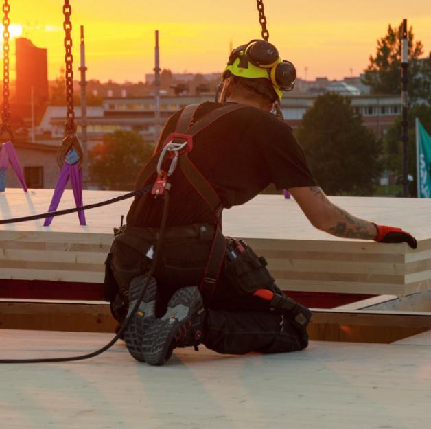 Construction worker with fall arrest harness guiding CLT panels onto a flat timber roof at sunset — flat timber roof construction and stratigraphy.