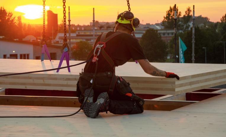 Construction worker with fall arrest harness guiding CLT panels onto a flat timber roof at sunset — flat timber roof construction and stratigraphy.